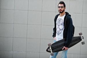 hombre árabe de estilo callejero en anteojos con longboard posado contra la pared gris. foto