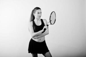 Black and white portrait of beautiful young woman player in sports clothes holding tennis racket while standing against white background. photo