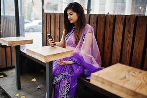 niña hindú india en el sari violeta tradicional sentada en la mesa de café con el teléfono móvil en las manos. foto