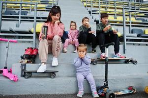 joven madre elegante con cuatro hijos sentados en el podio deportivo en el estadio, come manzana y bebe agua. la familia pasa tiempo libre al aire libre con scooters y patines. foto
