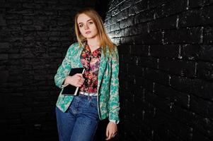 Stylish blonde girl in jacket and jeans with notebook diary at hands against brick black wall at studio. photo