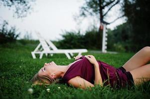 Blonde sensual woman in red marsala dress lying in the green grass. photo