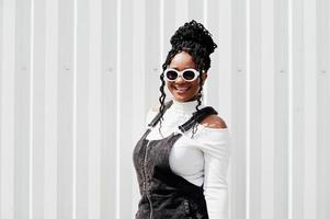 African woman in denim overalls skirt, white sunglasses posed against white steel wall. photo