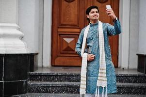 hombre indio vestido con ropa tradicional con bufanda blanca posada al aire libre contra la puerta del edificio con teléfono móvil a mano y haciendo selfie. foto