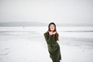 ropa de niña divertida en sudadera verde larga, jeans y sombreros negros, en el lago congelado en el día de invierno. foto