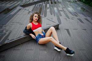 Sexy curly model girl in red top, jeans denim shorts, leather jacket and sneakers posed on the floor. photo