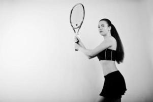 Black and white portrait of beautiful young woman player in sports clothes holding tennis racket while standing against white background. photo