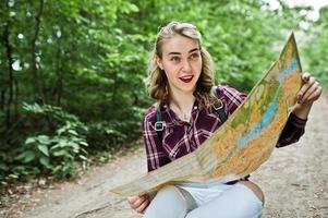 retrato de una joven y hermosa rubia positiva sentada en el suelo con un mapa en las manos en el bosque. foto