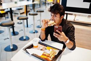 Stylish and funny indian man sitting at fast food cafe and eating hamburger and making selfie by phone. photo