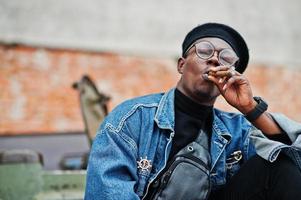 African american man in jeans jacket, beret and eyeglasses, smoking cigar and posed against btr military armored vehicle. photo