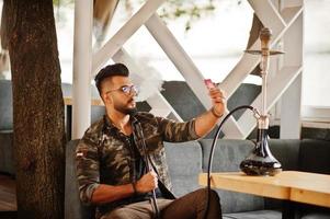 hombre árabe de barba elegante con gafas y chaqueta militar fumando narguile en el bar de la calle. modelo árabe descansando y tomando selfie en el teléfono. foto