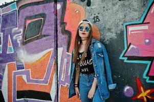 Stylish casual hipster girl in cap, sunglasses and jeans wear, listening music from headphones of mobile phone against large graffiti wall. photo