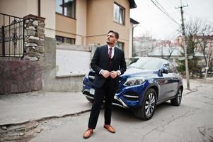 Rich indian businessman in formal wear standing against business suv car and his large mansion house. photo