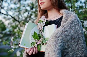 joven morena en tela escocesa contra el árbol de flores de primavera y sosteniendo el libro en las manos. foto