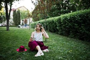 Fashionable and beautiful blonde model girl in stylish red velvet velour skirt, white blouse and hat, sitting on green grass at park with phone and earphones. photo