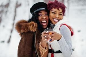 Two african american womans in sheepskin and fur coat posed at winter day against snowy background with cups of coffee. photo