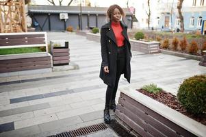 Portrait of a curly haired african woman wearing fashionable black coat and red turtleneck posing outdoor. photo