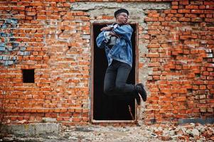 African american man in jeans jacket, beret and eyeglasses against brick wall jump at abandoned roof. photo