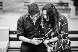 Indian couple posed outdoor, sitting on bench together and looking at mobile phone. photo