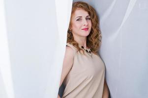 Curly hair girl on beige dress posed at white curtains. photo