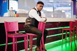 Young handsome african man wearing white shirt, black vest and bow tie posed against bar counter at night club. photo