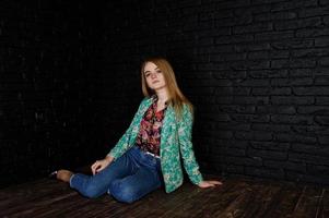 Stylish blonde girl in jacket and jeans against brick black wall at studio. photo