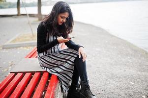 Portrait of young beautiful indian or south asian teenage girl in dress sitting on bench with mobile phone. photo