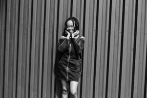 African woman in denim overalls skirt, eyeglasses posed against green steel wall. photo