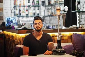 Stylish beard arabian man in glasses and black t-shirt smoking hookah indoor bar. Arab model having rest. photo