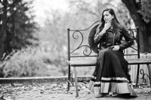 linda chica india con vestido de sari negro y chaqueta de cuero posada al aire libre en la calle de otoño y sentada en un banco. foto