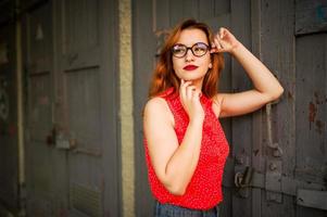 Attractive redhaired woman in eyeglasses, wear on red blouse and jeans skirt posing. photo