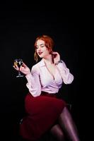 Gorgeous red haired girl in pink blouse and red skirt with glass of wine at hand isolated on black. photo