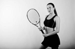 Black and white portrait of beautiful young woman player in sports clothes holding tennis racket while standing against white background. photo