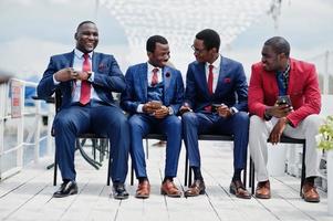 grupo de cuatro hombres afroamericanos felices y exitosos en traje. hombres de negocios negros ricos sentados en sillas mirando teléfonos móviles. foto