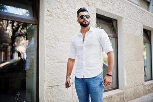 Stylish tall arabian man model in white shirt, jeans and sunglasses posed at street of city. Beard attractive arab guy. photo