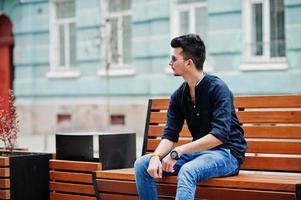 Stylish indian model man in casual clothes, black shirt and sunglasses posed outdoor at street of India and sitting on bench. photo