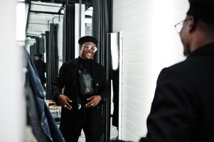 Stylish casual african american man at black beret and overcoat with waist bag at fitting room clothes store, looking on mirror. photo