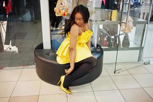 elegante mujer afroamericana vestida de amarillo sentada en una bocanada contra vitrinas en el centro comercial. foto