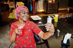Stylish african woman in red shirt and hat posed indoor cafe, drinking strawberry lemonade and making selfie on phone. photo
