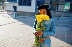 mujeres afroamericanas de moda con estilo en jeans y boina negra con ramo de flores amarillas posadas al aire libre en un día soleado contra un edificio azul moderno. foto