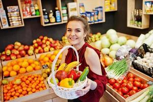 Girl in red holding different fruit and vegetables at basket on fruits store. photo