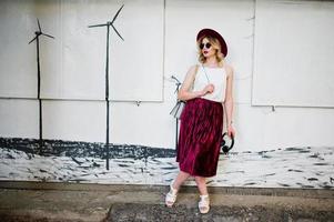 Fashionable and beautiful blonde model girl in stylish red velvet velour skirt, white blouse, hat and sunglasses, posed with phone and earphones. photo