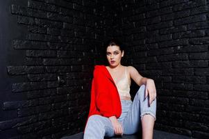 Stylish brunette girl on red jacket against studio black brick wall. photo