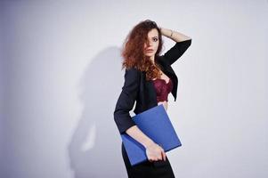 Brunette curly haired girl in black office jacket with skirt, on bra with folder of documentts at hands on studio against white background. Sexy businesswoman. photo