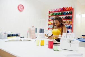 Young african dressmaker woman sews clothes on sewing machine at tailor office. Black seamstress women. photo