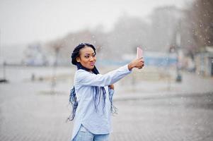 Stylish african american girl with dreads holding mobile phone at hand and making selfie, outdoor with snowy weather. photo