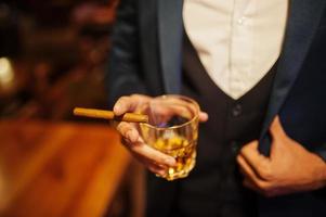 Close up hand of handsome well-dressed arabian man with glass of whiskey and cigar posed at pub. photo