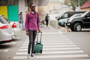 African american man in checkered shirt, sunglasses and earphones with suitcase and backpack. Black man traveler walking on crosswalk. photo