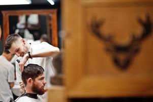 Young bearded man getting haircut by hairdresser while sitting in chair at barbershop. Barber soul. photo