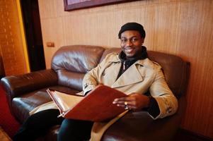 Handsome african american man posing  inside room with sunlight shadows in black hat and beige coat. photo